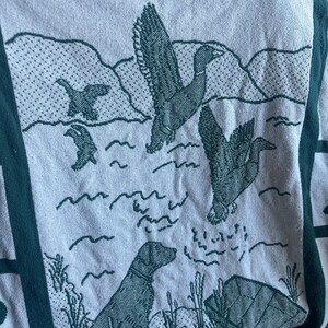 Pu&ograve; includere: Una coperta di colore chiaro con un bordo verde e una scena naturale. Il design presenta anatre in volo sull'acqua, un cane e canne. La coperta ha un motivo ripetuto.