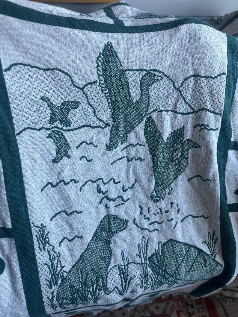 Pu&ograve; includere: Una coperta di colore chiaro con un bordo verde e una scena naturale. Il design presenta anatre in volo sull'acqua, un cane e canne. La coperta ha un motivo ripetuto.