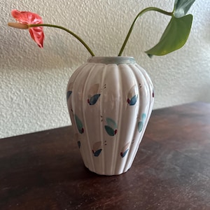 May include: A white ceramic vase with a ribbed design and a light blue rim. The vase is decorated with blue, teal, and red abstract shapes. It holds a red anthurium flower and a green leaf.
