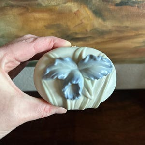 May include: An oval-shaped ceramic trinket box with a matte cream-coloured finish. The lid features a raised, three-dimensional sculpted flower with soft blue petals and green stems, providing a textured and detailed decorative accent to the piece.