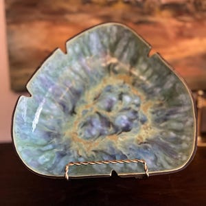 May include: A decorative ceramic dish with a unique, organic shape. The dish features a blend of blue, green, and gold hues, with a glossy finish. It is displayed on a gold-colored metal stand, showcasing its artistic design. The dish's edges are dark.