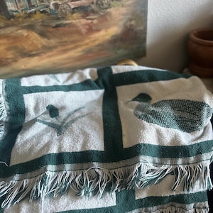 Pu&ograve; includere: Una coperta piegata bianca e verde con un bordo a frange. La coperta presenta un motivo ripetuto di bordi verde scuro e disegni stilizzati di uccelli. La coperta &egrave; drappeggiata su una superficie, con un dipinto sullo sfondo.