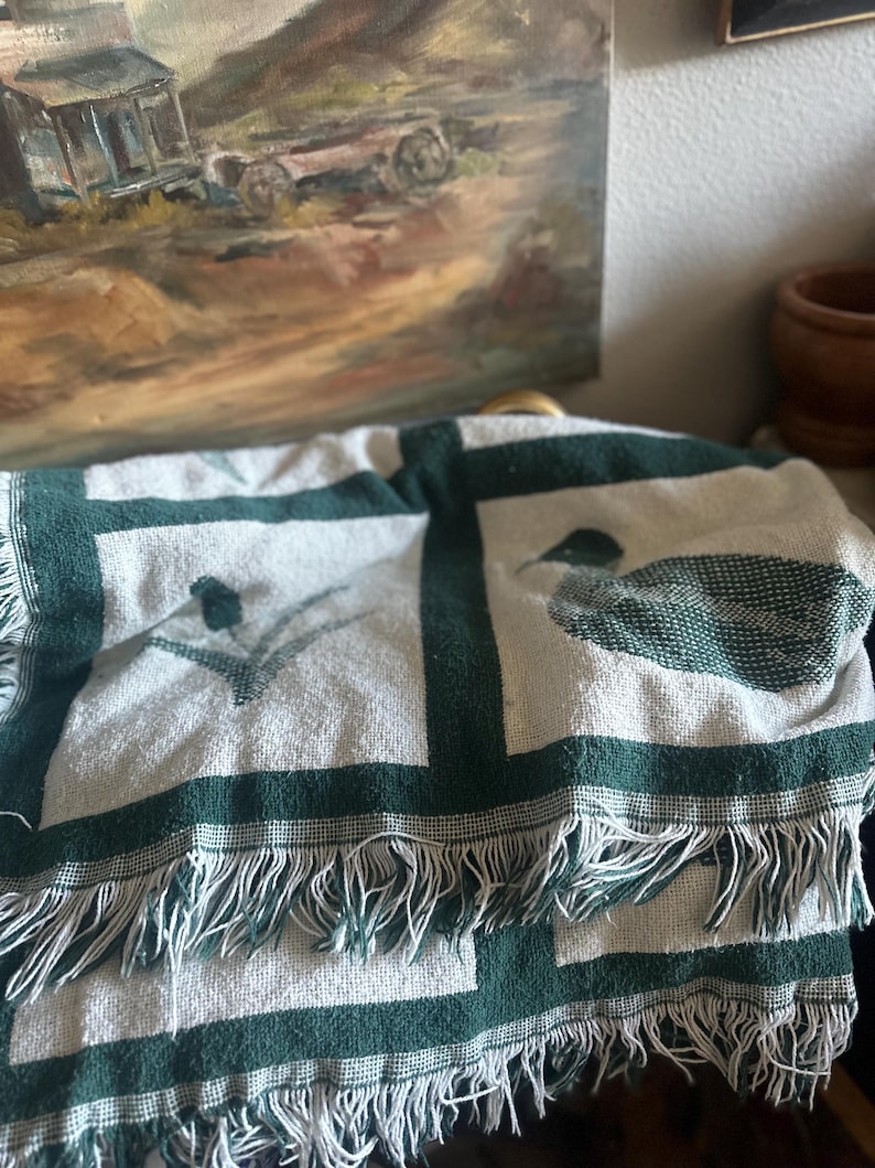 Pu&ograve; includere: Una coperta piegata bianca e verde con un bordo a frange. La coperta presenta un motivo ripetuto di bordi verde scuro e disegni stilizzati di uccelli. La coperta &egrave; drappeggiata su una superficie, con un dipinto sullo sfondo.