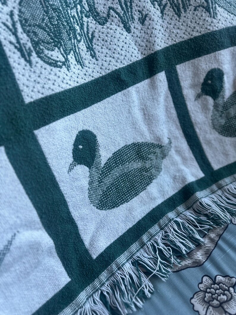Pu&ograve; includere: Una coperta tessuta con un motivo ripetuto di anatre in verde scuro su sfondo grigio chiaro. La coperta ha un bordo verde scuro e frange sul bordo inferiore. Le anatre sono raffigurate in modo dettagliato.