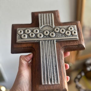 May include: A wall cross featuring a dark brown wooden base with a central silver-toned metal inlay. The metal piece displays a horizontal band of circular motifs above a vertical section of parallel raised lines, creating a textured contrast.
