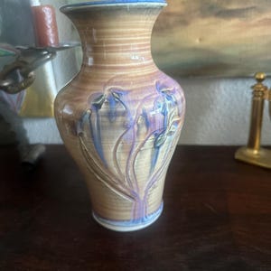 May include: A ceramic vase with a flared rim and a tapered neck, featuring a tan and brown glazed finish with a subtle crackle texture. The body is decorated with embossed, stylized blue and purple floral motifs that wrap around the exterior.