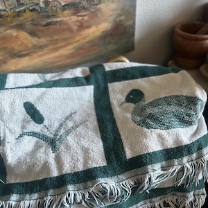 Pu&ograve; includere: Una coperta bianca e verde con un motivo di anatra e giunchi. La coperta ha un bordo con frange e un bordo verde scuro. La coperta &egrave; piegata e sembra essere fatta di un materiale tessuto.