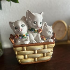 May include: A ceramic figurine featuring three white kittens in a woven basket. The kittens have green eyes and are adorned with floral collars. The basket is painted in shades of yellow and brown, creating a charming decorative piece.