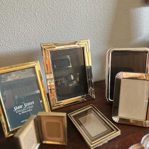 May include: A collection of gold-toned picture frames in various sizes and styles. Some frames have a rectangular shape, while others are square or have rounded edges. One frame displays the text "State Street Collection by Hilburnes."