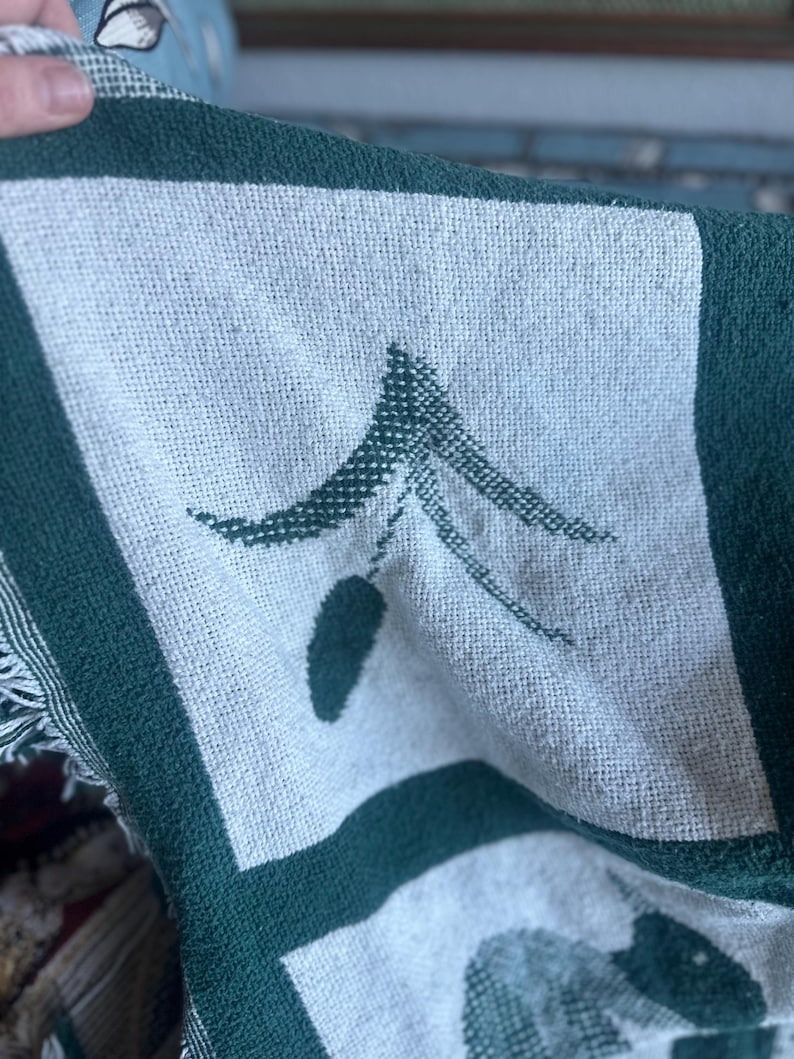 Pu&ograve; includere: Primo piano di una coperta tessuta con un motivo ripetuto di quadrati verdi e bianchi. Ogni quadrato presenta un design ad albero stilizzato. La coperta ha un bordo verde scuro e frange.