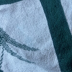 Pu&ograve; includere: Primo piano di una coperta tessuta bianca e verde. La coperta ha un centro bianco con un bordo verde e un disegno di palma verde nell'angolo. La trama della coperta &egrave; visibile.