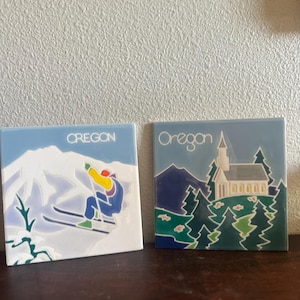 May include: Two square ceramic tiles with the word "Oregon" printed on them. One tile depicts a skier on a snowy mountain, the other shows a church and trees. The tiles are blue and white.