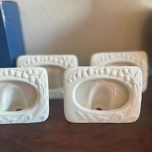 May include: Four white ceramic picture frames with oval apertures and floral detailing. The frames are rectangular with curved tops and bottoms. They are arranged on a dark wooden surface.