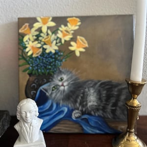 May include: A painting of a gray cat lying on a blue fabric with a vase of yellow daffodils in the background. A white bust of a person is in the foreground. A gold candle holder with a white candle is on the right side of the image.