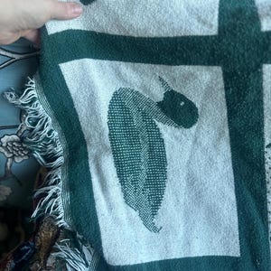 Pu&ograve; includere: Una coperta verde e bianca con un disegno di cavallo. La coperta ha un bordo con frange e un motivo ripetuto di cavalli all'interno di quadrati verdi. La coperta &egrave; realizzata in un materiale morbido e tessuto.