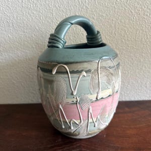May include: A ceramic vase with a teal handle and rim, featuring a pink and gray body with white, textured, abstract designs. The vase has a rounded shape and a looped handle.