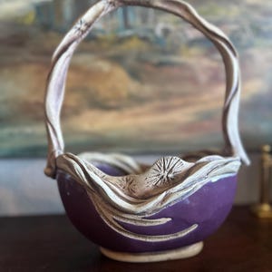 May include: A ceramic basket featuring a deep purple bowl and a light beige, textured rim and handle. The rim is decorated with carved starburst patterns and flowing, ribbon-like edges that extend from the base of the handle to the bowl.