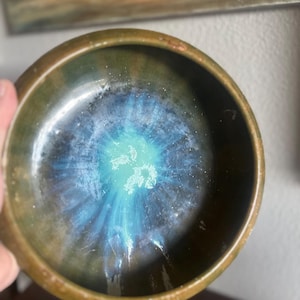 May include: A small, round ceramic bowl with a dark green exterior and a vibrant, blue and turquoise interior. The interior glaze has a unique, swirling pattern, creating a celestial effect. The bowl's rim is a darker shade of green.