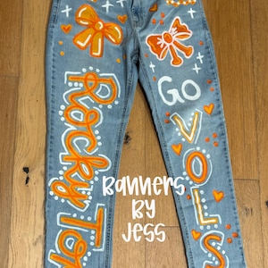 May include: Light blue denim jeans with hand-painted orange and white accents. Features include orange bows, hearts, and the words "Rocky Top" and "Go Vols". The text "Banners by Jess" is also visible.