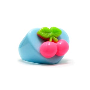 May include: A light blue ring with a cherry design. The ring features two pink cherries and green leaves on top. The ring is a fun, playful accessory.