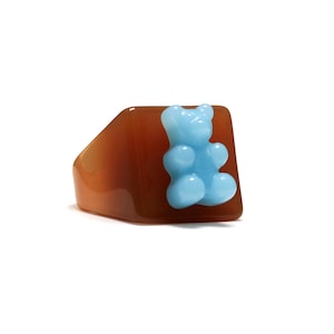 Pode incluir: Um anel robusto com uma banda quadrada de cor castanha translúcida e superfície lisa. Na parte frontal central da banda, encontra-se uma pequena figura de urso de goma azul-claro opaco com acabamento brilhante, criando um contraste visual.