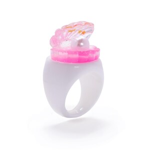 May include: A white ring with a pink seashell design. The seashell is open, revealing a pearl. The ring has a smooth, glossy finish, and the seashell has glitter and star accents.