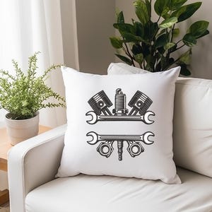 May include: White square throw pillow with a black graphic of automotive tools, including pistons, a spark plug, and wrenches. The pillow is on a white sofa, with a small wooden table and a potted plant in the background.