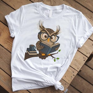 May include: White t-shirt featuring an embroidered owl wearing glasses and reading a book. The owl is perched on a branch with a stack of books and a coffee cup. The design is in shades of brown, blue, and green.