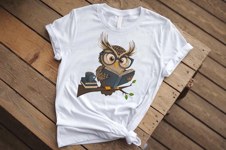 May include: White t-shirt featuring an embroidered owl wearing glasses and reading a book. The owl is perched on a branch with a stack of books and a coffee cup. The design is in shades of brown, blue, and green.