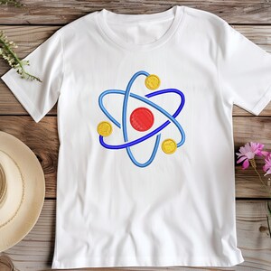 K&ouml;nnte beinhalten: Wei&szlig;es T-Shirt mit einem gestickten Atom-Design. Das Atom hat eine rote Mitte, blaue Umlaufbahnen und gelbe kreisf&ouml;rmige Akzente. Das Shirt wird auf einer Holzoberfl&auml;che mit einem Strohhut und Blumen pr&auml;sentiert.