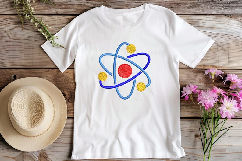 K&ouml;nnte beinhalten: Wei&szlig;es T-Shirt mit einem gestickten Atom-Design. Das Atom hat eine rote Mitte, blaue Umlaufbahnen und gelbe kreisf&ouml;rmige Akzente. Das Shirt wird auf einer Holzoberfl&auml;che mit einem Strohhut und Blumen pr&auml;sentiert.