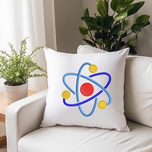 K&ouml;nnte beinhalten: Wei&szlig;es quadratisches Kissen mit einem gestickten Atom-Design. Das Atom hat eine rote Mitte, blaue Umlaufbahnen und drei gelbe kreisf&ouml;rmige Elemente. Das Kissen liegt auf einem wei&szlig;en Sofa in der N&auml;he eines kleinen Holztisches mit einer Topfpflanze.