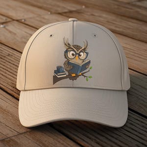 May include: A light beige baseball cap featuring an embroidered owl wearing glasses and reading a book. The owl is perched on a branch with a small stack of books and a coffee cup. The cap is on a wooden surface.