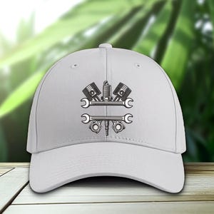 May include: A white baseball cap featuring a black and gray graphic of automotive tools, including pistons, a spark plug, and wrenches. The design is centered on the front of the cap, suitable for mechanics and car enthusiasts.