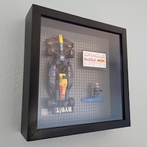 May include: A black framed shadow box with a Lego Formula 1 race car, a Lego driver figure, and a gray Lego baseplate. The car is black and yellow with the Oracle Red Bull Racing logo on the side. The baseplate has the words "BYBIT" printed on it.