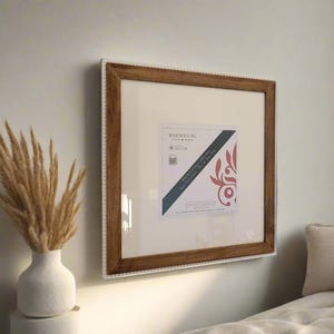 May include: Square wooden picture frame with a white mat and a decorative border of small white beads. The frame is hanging on a light-colored wall. The frame contains a printed image with text and a floral design.