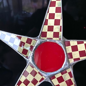 May include: A star-shaped decorative object with a red center and a checkered pattern in red and gold. The star's points have a silver border, and the background is black.