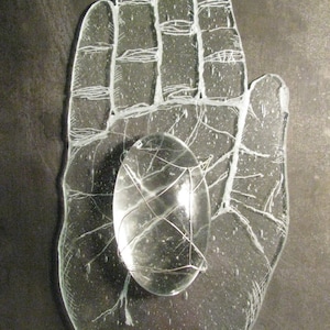 May include: A clear glass hand sculpture with a smaller, round, clear glass orb in the palm. The hand is detailed with lines and textures.