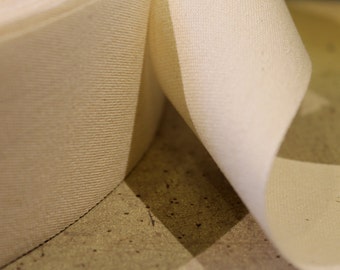 May include: Close-up of a roll of off-white fabric. The fabric has a textured weave and is partially unrolled, revealing the inside. The lighting creates shadows and highlights the texture of the material. The fabric appears to be cotton or a similar natural fiber.