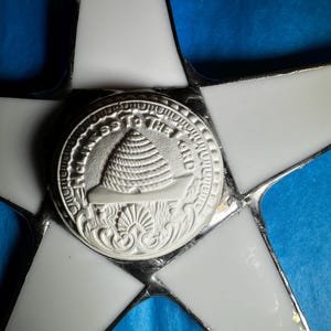 May include: A white star-shaped stained glass window with a silver-colored circular medallion in the center. The medallion features a raised image of a ship with the text "In God We Trust" around the edge.