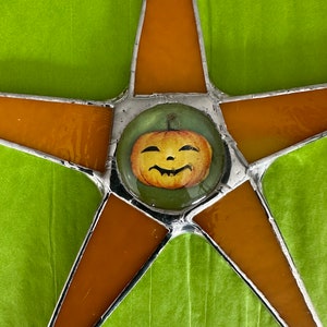 May include: A stained glass star with orange and clear panels, and a smiling jack-o'-lantern in the center. The star is outlined in silver and set against a green background, perfect for Halloween decor.