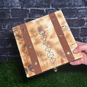 May include: Wooden box with a decorative design, featuring a sword, dice, and geometric shapes. The box has brown leather straps and brass rivets. The box is held by a hand.