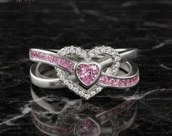 May include: A silver ring with a heart-shaped design. The ring features a pink heart-shaped center stone, surrounded by white gemstones and pink gemstones. The band has an infinity symbol design.