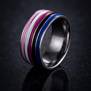 Op de afbeelding: Een brede metalen ring met vijf afzonderlijke verzonken groeven, elk gevuld met een emaille inleg in een andere kleur. De kleuren zijn lichtroze, wit, magenta, zwart en levendig koningsblauw, wat zorgt voor een gestreept effect op de ring.