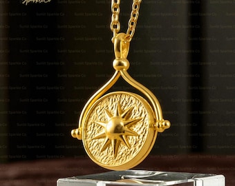 May include: A gold-colored pendant necklace featuring a textured sunburst design. The pendant is circular and rotates within a gold frame, suspended from a delicate chain. The necklace is displayed on a clear stand.