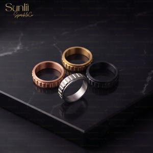 Op de afbeelding: Vier metalen ringen met een getextureerde, tandwielachtige buitenrand. De set bevat een roségouden, gouden, zilveren en zwarte ring. Elke ring heeft een gladde binnenband en een gegroefd, industrieel ogend ontwerp aan de buitenzijde.