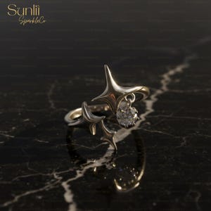 Pode incluir: Um anel de prata com um design de estrela. O anel apresenta dois detalhes em forma de estrela e uma pedra preciosa transparente pendurada. O anel está sobre um fundo escuro com o texto "Sunlii SparkleCo" no canto superior esquerdo.