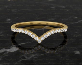 May include: A gold-colored, V-shaped ring with a row of small, clear gemstones set along the top. The ring is displayed on a dark, reflective surface, highlighting its sparkle and design. The ring is a piece of jewelry.