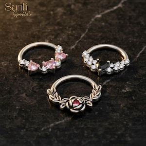 Puede incluir: Tres anillos de nariz plateados con diferentes diseños. Uno presenta gemas rosas en forma de corazón y piedras transparentes. Otro tiene una gema negra rodeada de piedras transparentes. El tercer anillo tiene un diseño de rosa y hojas con un centro rojo.