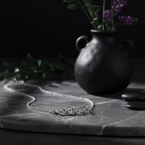 Puede incluir: Un collar plateado con un colgante de doble hélice de ADN. El collar se exhibe sobre una superficie de mármol gris, con un jarrón negro y flores moradas en el fondo. Es una delicada pieza de joyería.